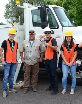 three students in hardhats and orange safety vests standing in front of a white truck with a man dressed in a khaki uniform
