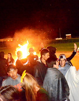 a group of students at night, many of them wrapped in blankets, standing in front of a blazing fire