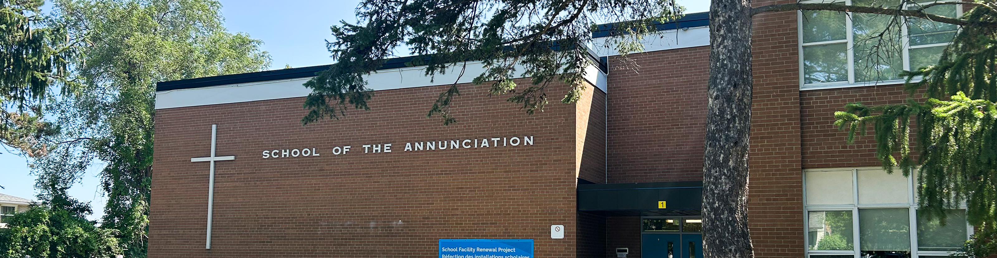 Photo of the front of the Annunication Catholic School building, showing a red brick building nestled beteen trees and bathed in sunlight.