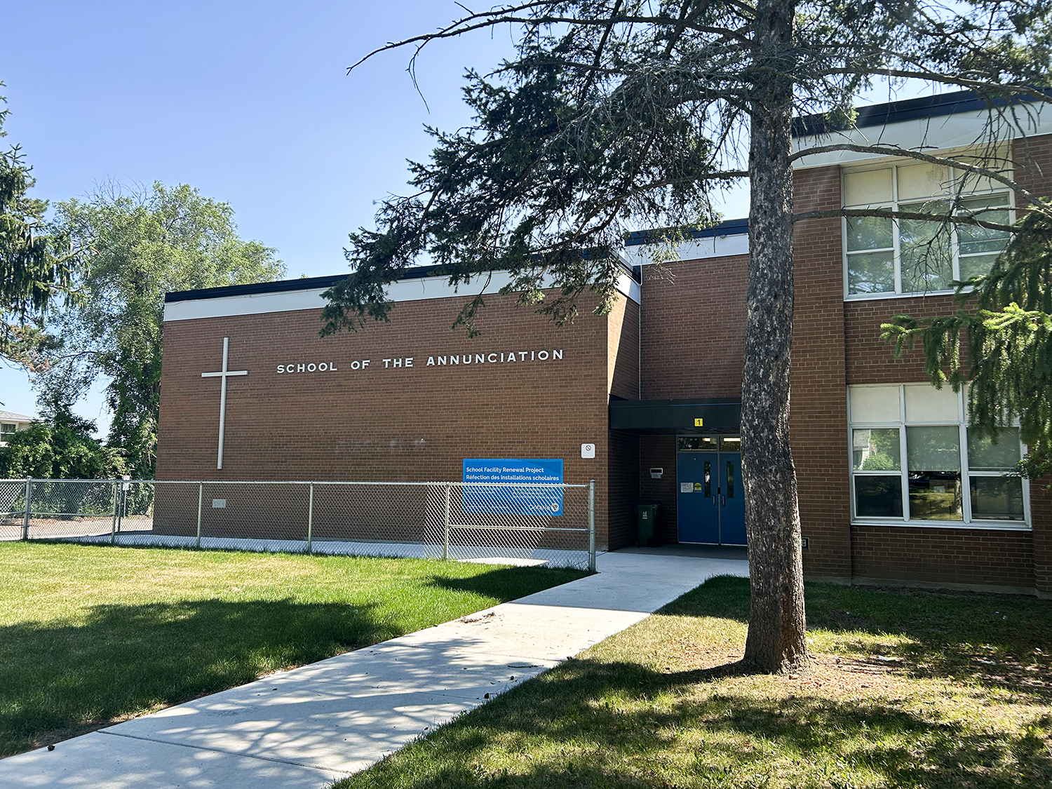 Photo of the front of the Annunication Catholic School building, showing a red brick building nestled beteen trees and bathed in sunlight.