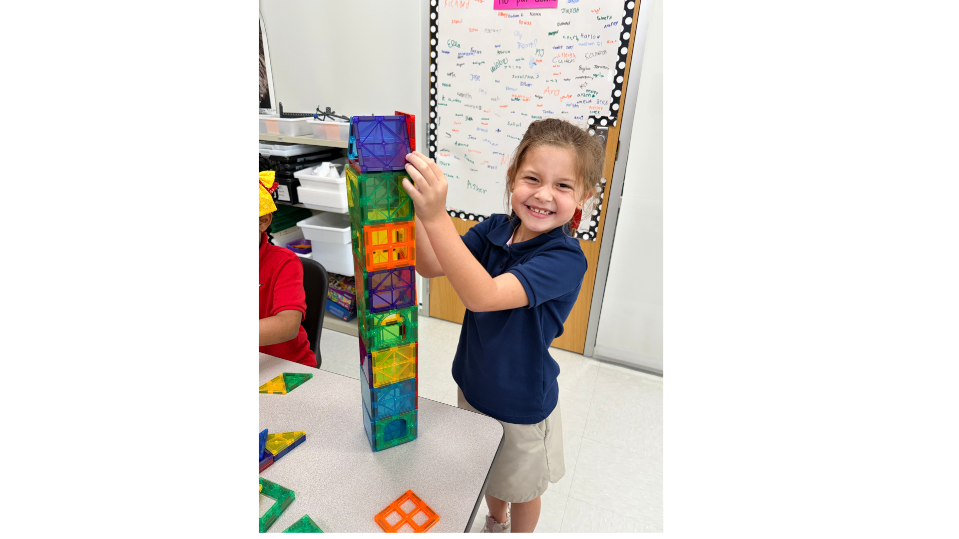 A child in a blue shirt and khaki shorts standing indoors, stacking colorful blocks on a table.