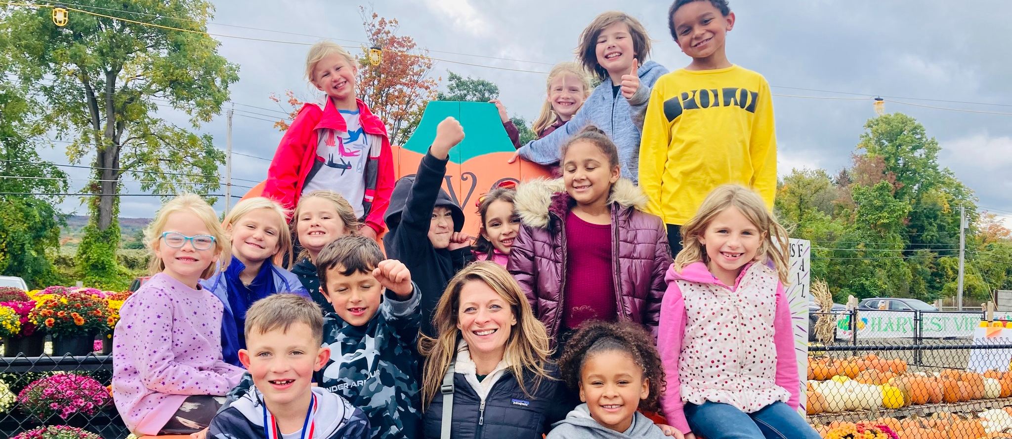 group photo of an elementary class with their teacher posing amongst mums, pumpkins, and other fall decor outside