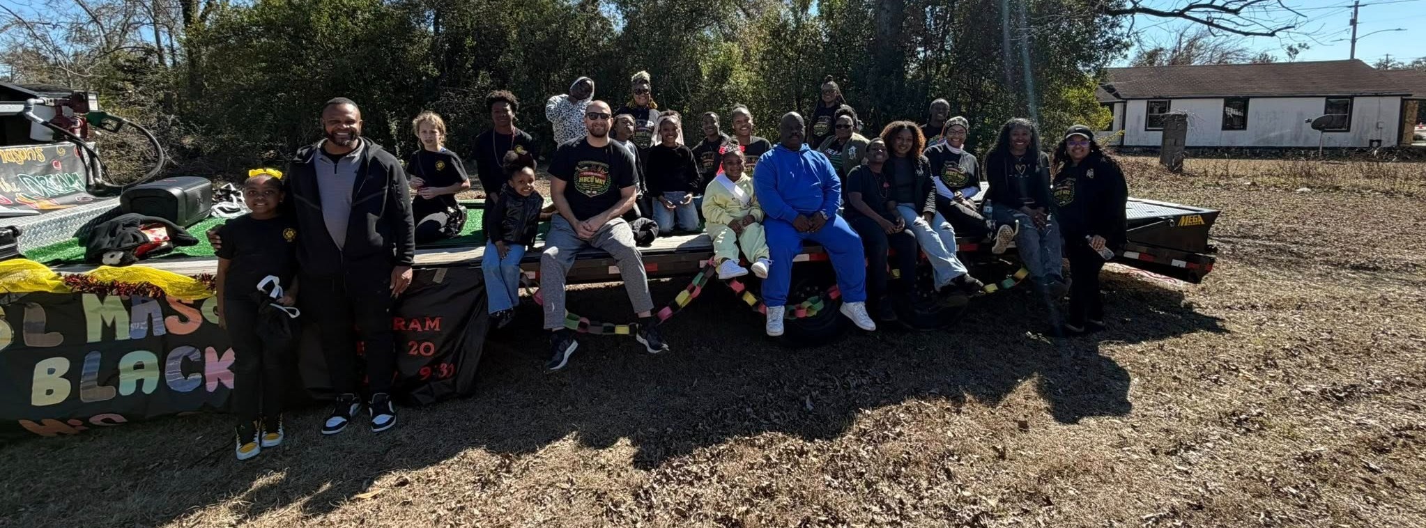 Group photo of about 20–25 people seated on a decorated flatbed trailer outdoors on a sunny day. The trailer is draped with yellow fabric and has linked red, green, and black chains. In the foreground a smiling Mr. Moody and young girl stand in front of the group.  A black banner shows partial text from the phrase "SL Mason Black History". Behind the group are dense green trees and some bare deciduous trees under a clear blue sky; a small white house with a dark roof is visible to the right. The ground appears dry and sparsely vegetated.
