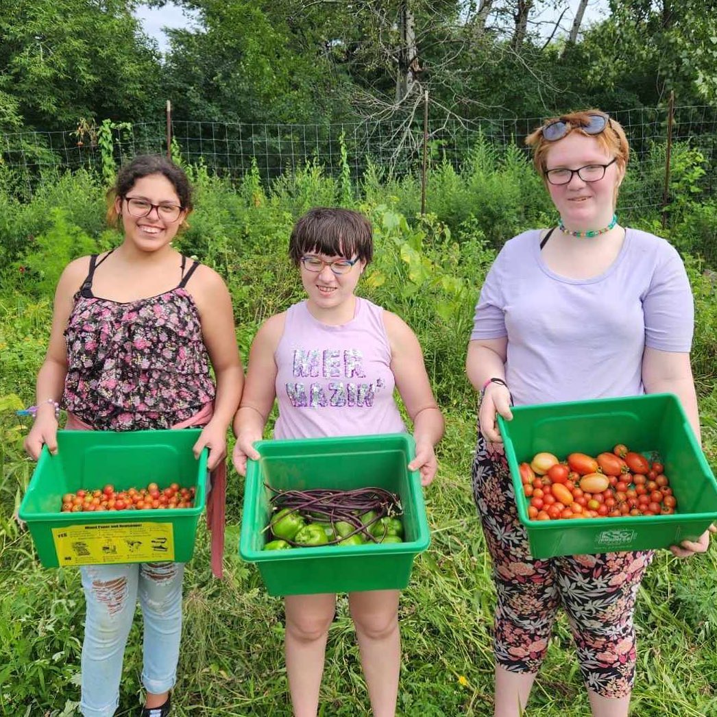 Three smiling girls stand holding their harvests of peppers, tomatoes, and green beans