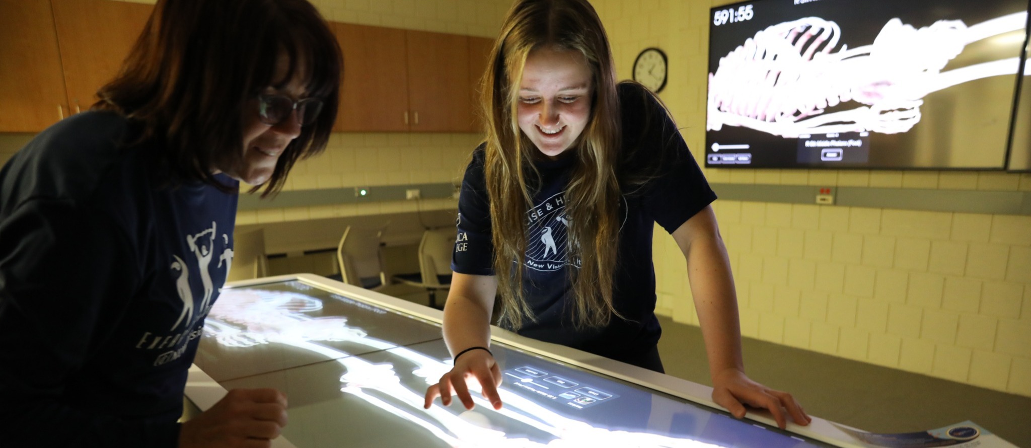 Two students interacting with a digital anatomy table displaying a human skeleton in a classroom lab.