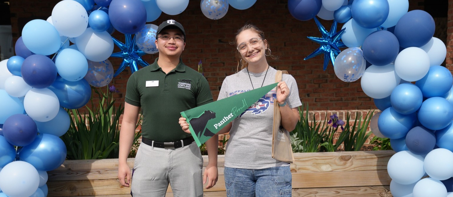 A student and a TC3 Admissions representative stand beneath a blue and white balloon arch during a college celebration event. One student holds a Tompkins Corltand green college pennant while both smile for a photo in front of a brick building and flower beds.