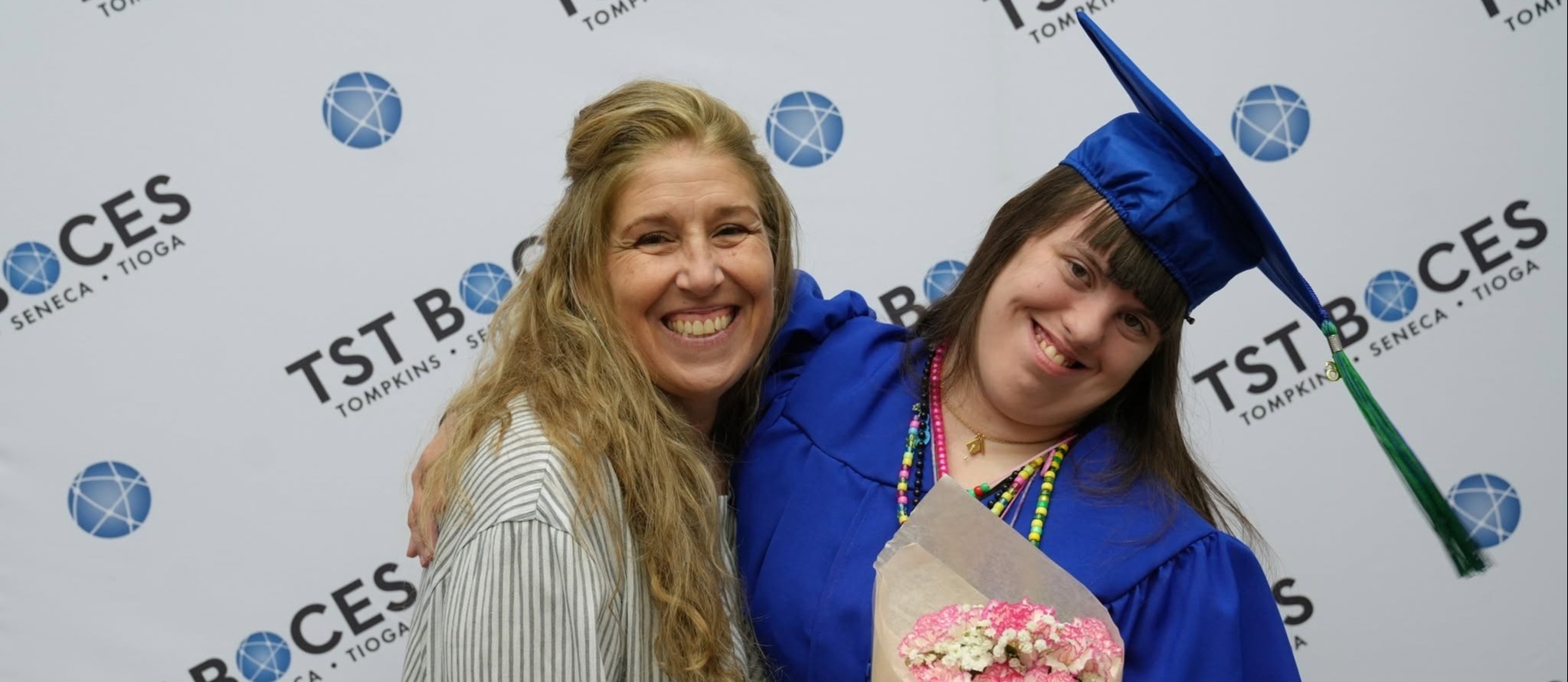 A smiling TST BOCES graduate in a blue cap and gown hugs a family member while holding a bouquet of pink flowers in front of a TST BOCES backdrop.