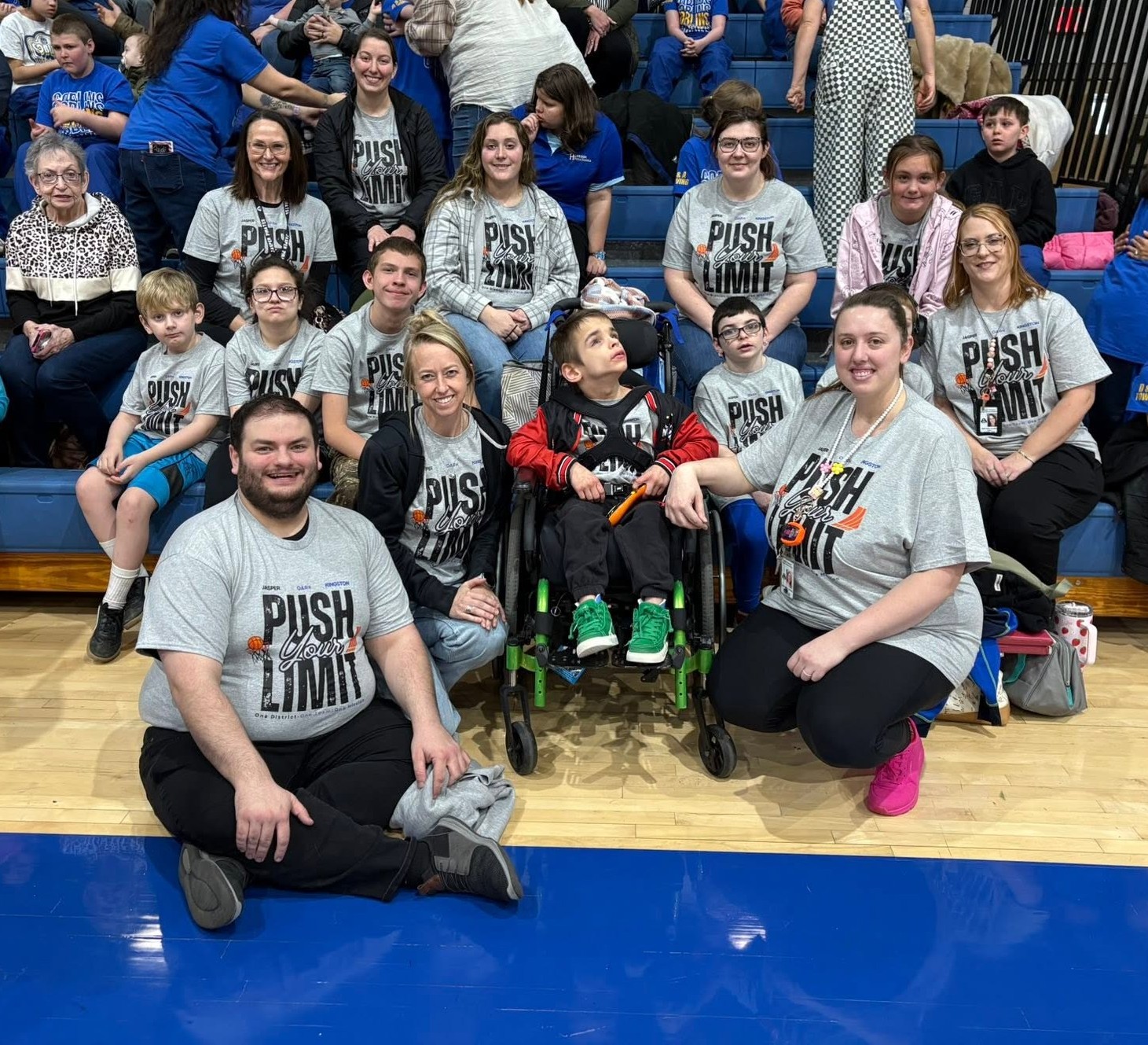 Group photo taken in a school gymnasium showing approximately 15 students and 10 adults seated and kneeling together on the gym floor and bleachers. Several individuals are wearing matching gray T-shirts that read “Push Your Limit.” A student using a wheelchair is positioned in the center of the group and is surrounded by classmates, staff members, and family supporters. Most participants are smiling and facing the camera. Blue bleachers filled with spectators are visible in the background. The image conveys inclusion, teamwork, support, and participation in a school athletic or special event.