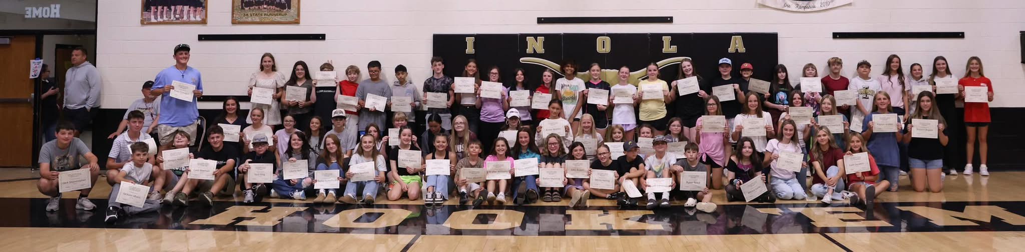 A group of students stands in a gymnasium, holding certificates, with a white wall and black border in the background.