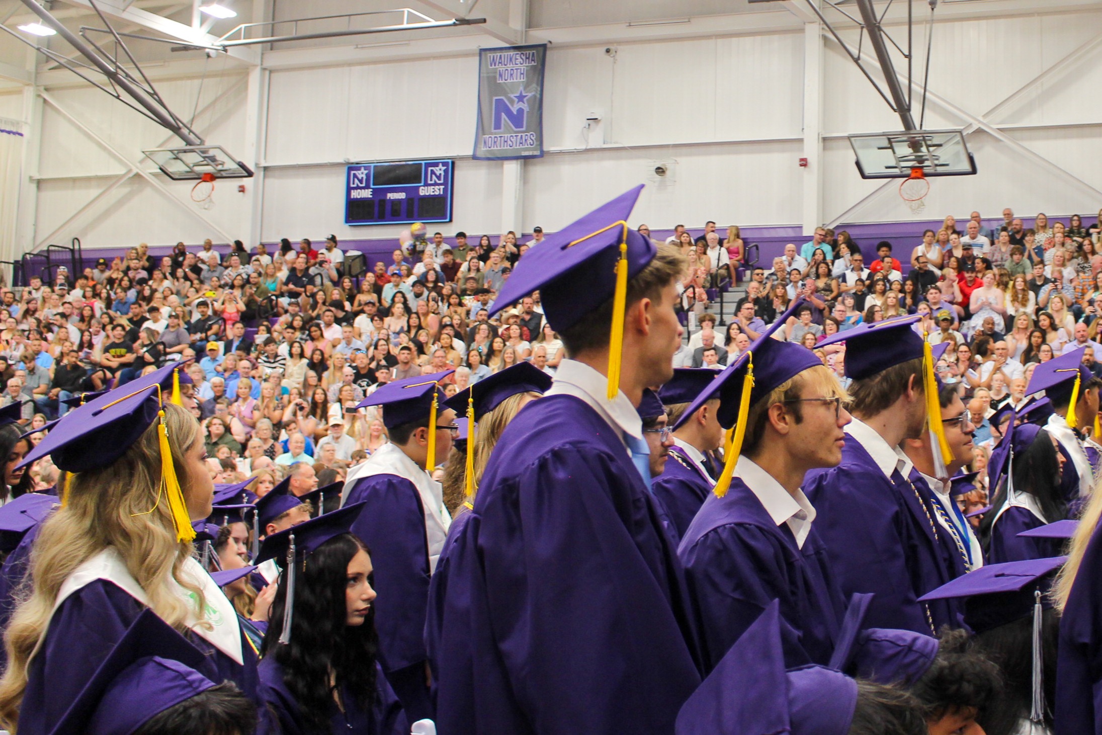 A group of people in caps and gowns are gathered in a large indoor venue, facing a large audience.