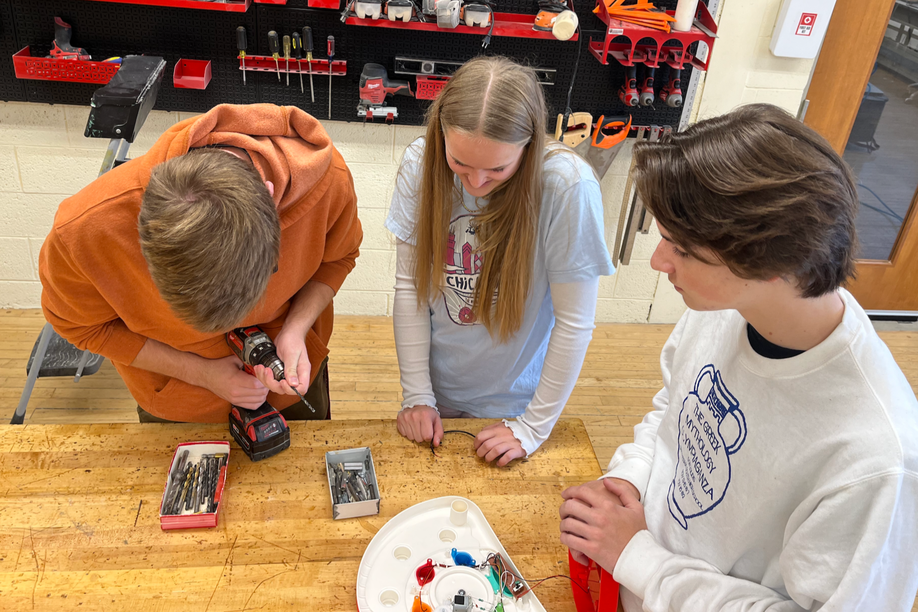 Three students gather around a table with tools, one drilling, one holding a tray of small objects, and another examining the objects.