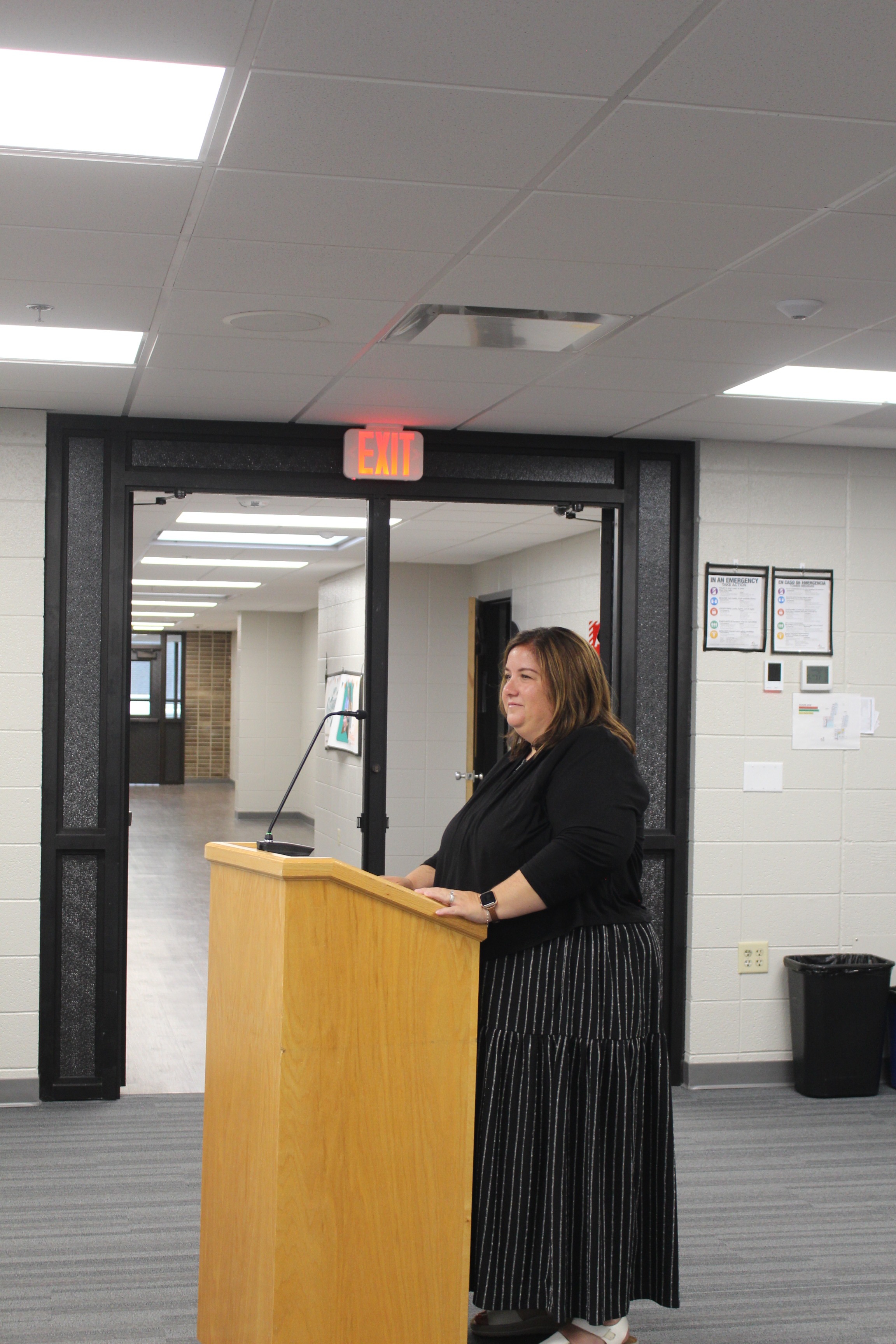 A woman stands at a podium, addressing the room. Behind her, a door leads to an open hallway.