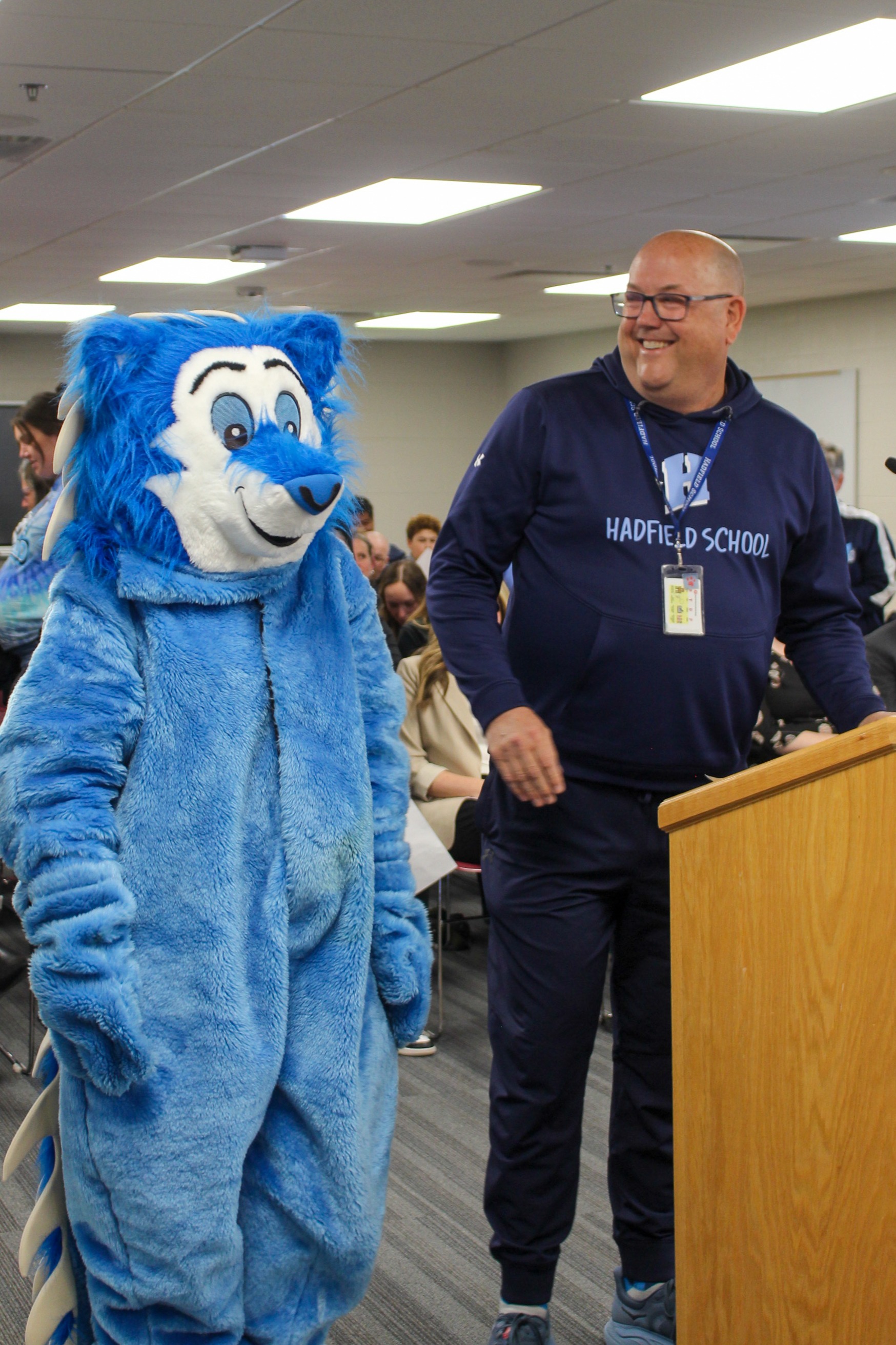 A man in a blue hoodie stands at a podium next to a blue mascot with a microphone.