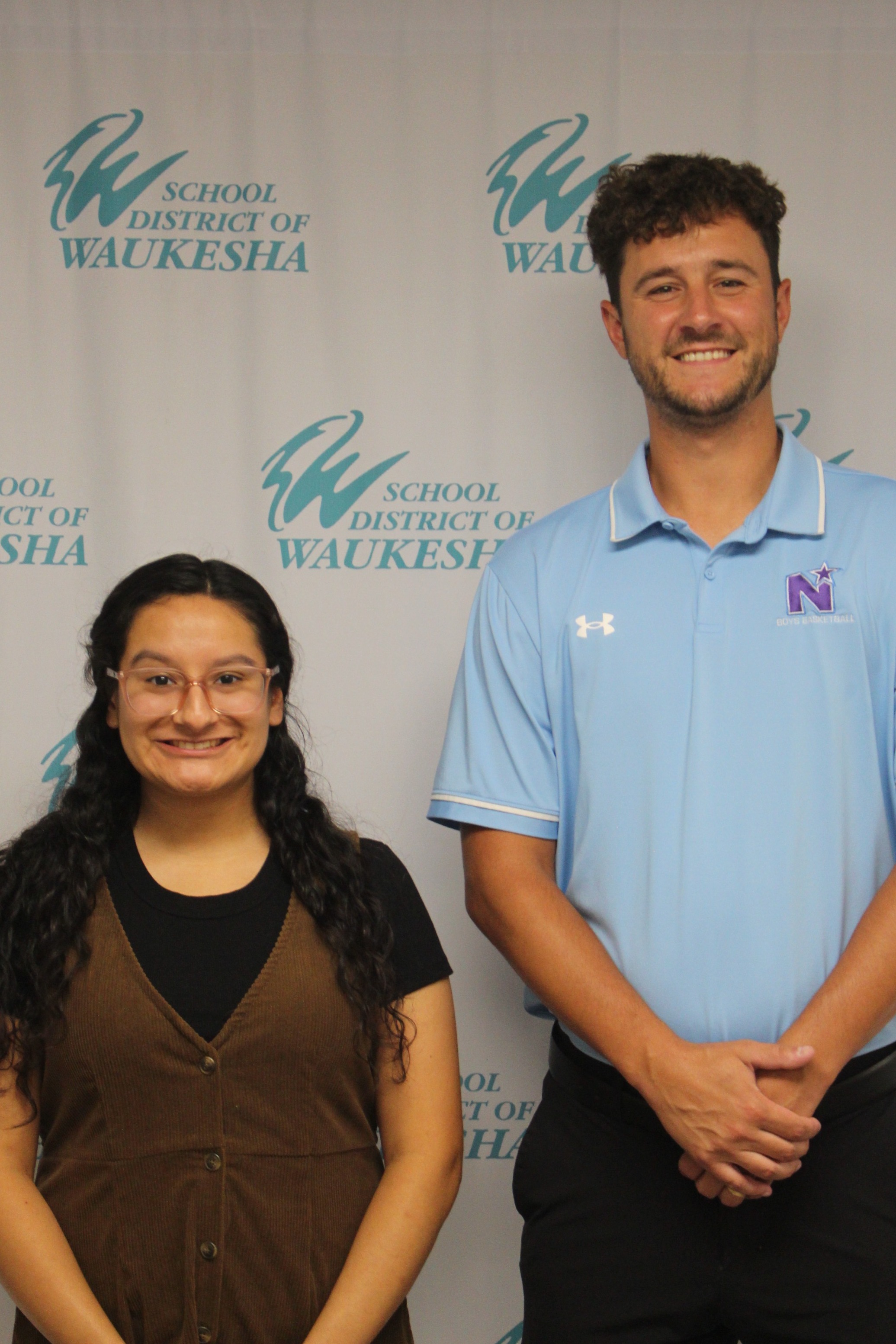 Two individuals stand in front of a white backdrop featuring the School District of Waukesha logo. A woman with glasses and a brown dress, and a man in a light blue polo.