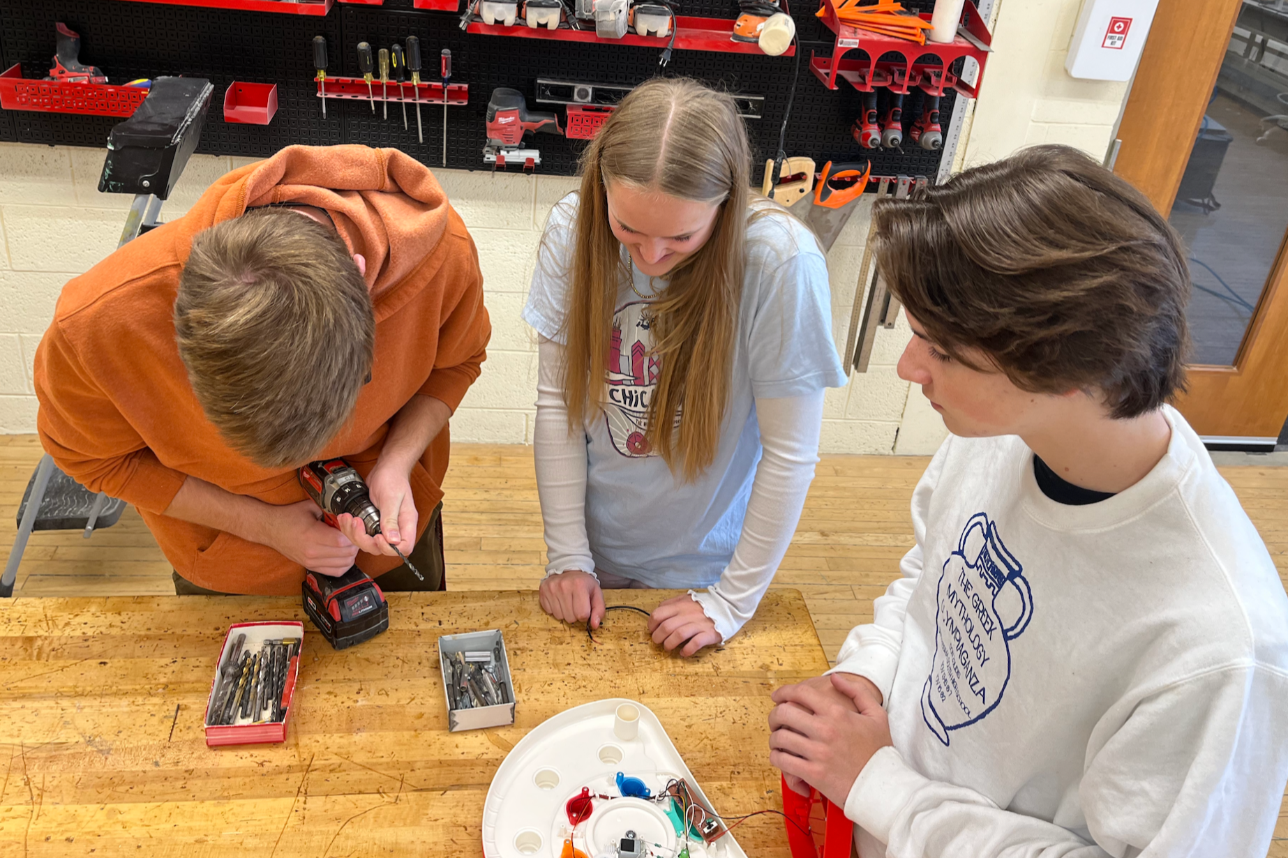 Three students collaborate at a workbench; a male using a drill, a girl with a white shirt, and another male. Behind them is a room with tools and shelves.
