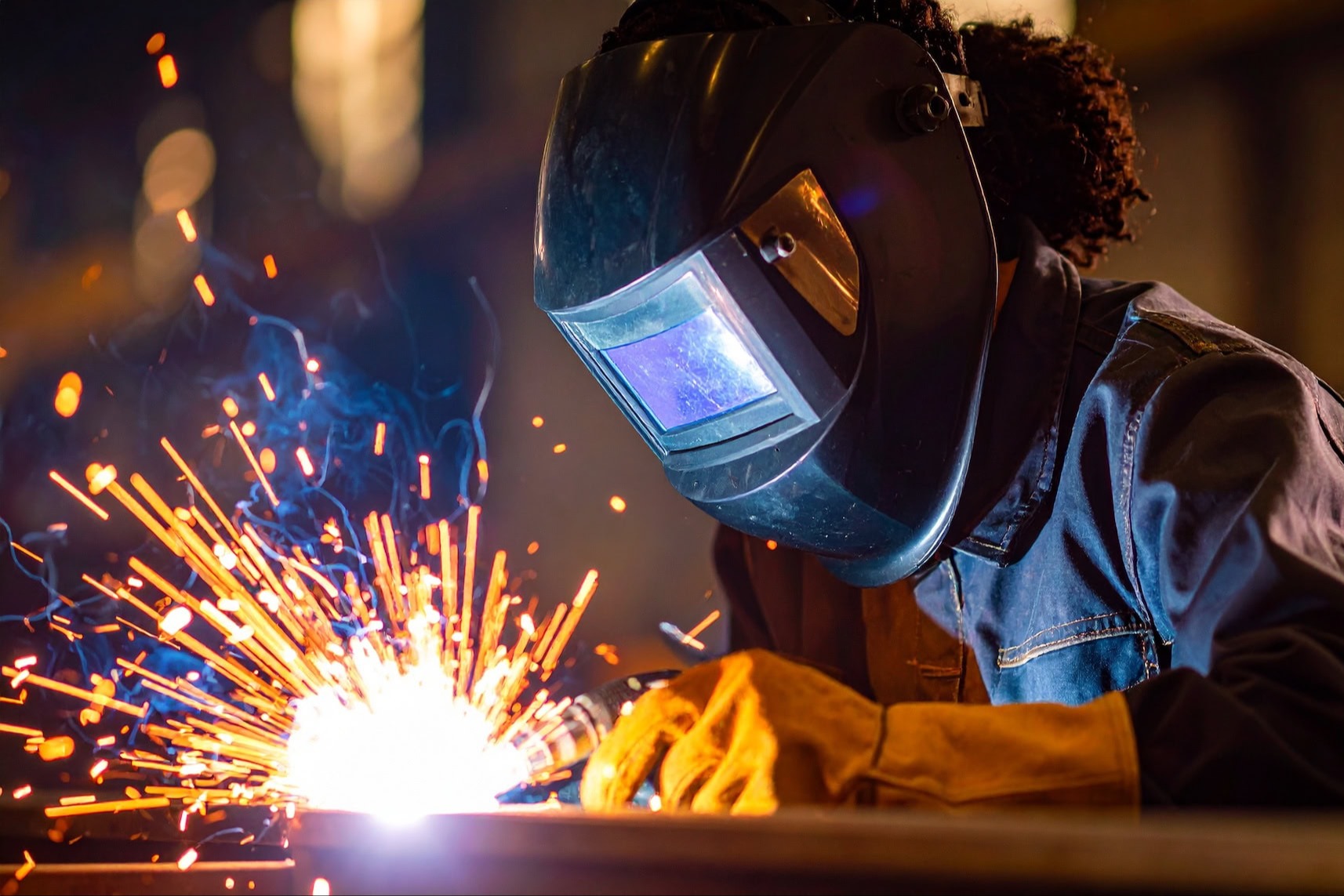 A person in a welding mask is welding metal, producing sparks. They wear gloves and a denim jacket.