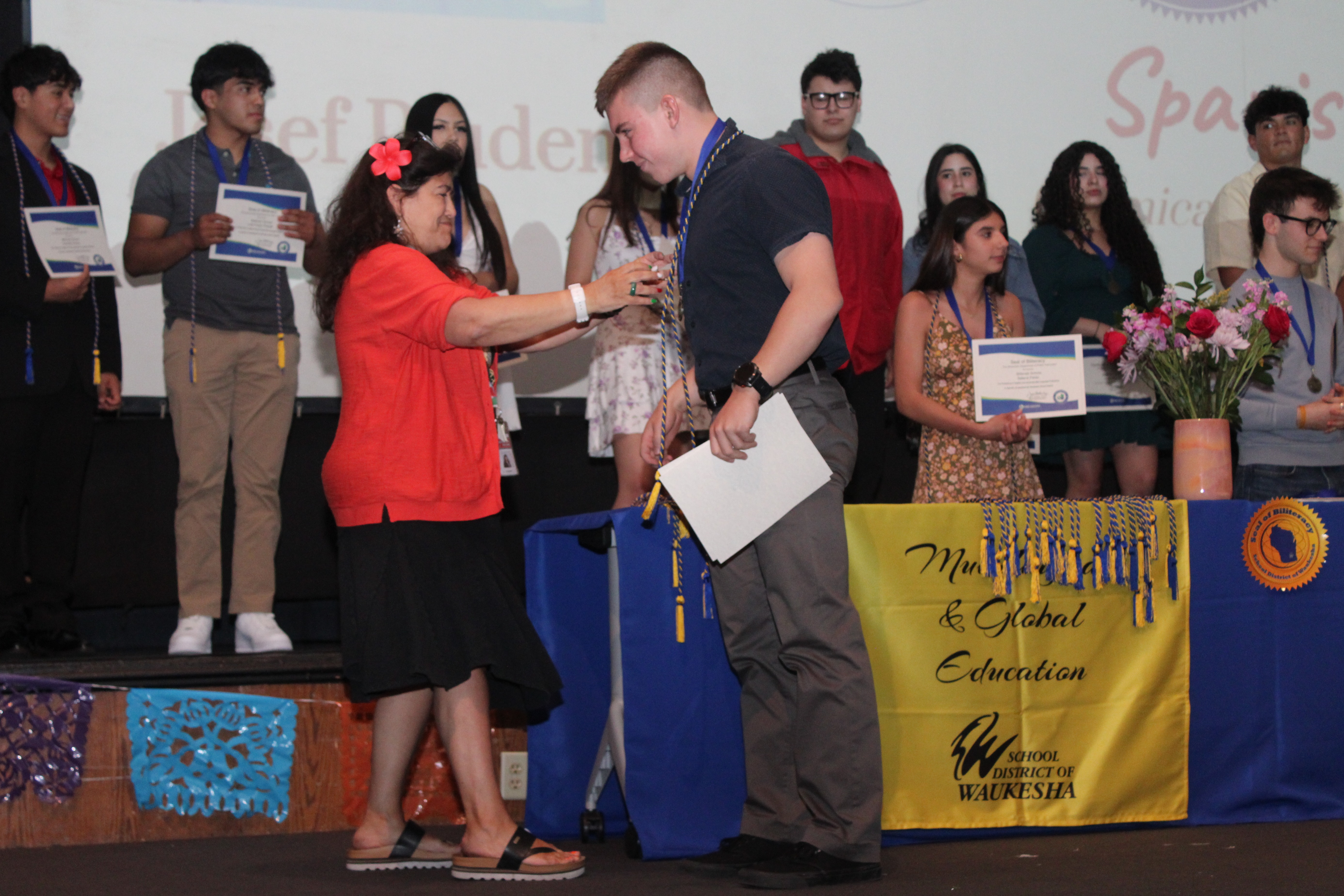 A woman on a stage gives an award to a man while others watch, some holding certificates.