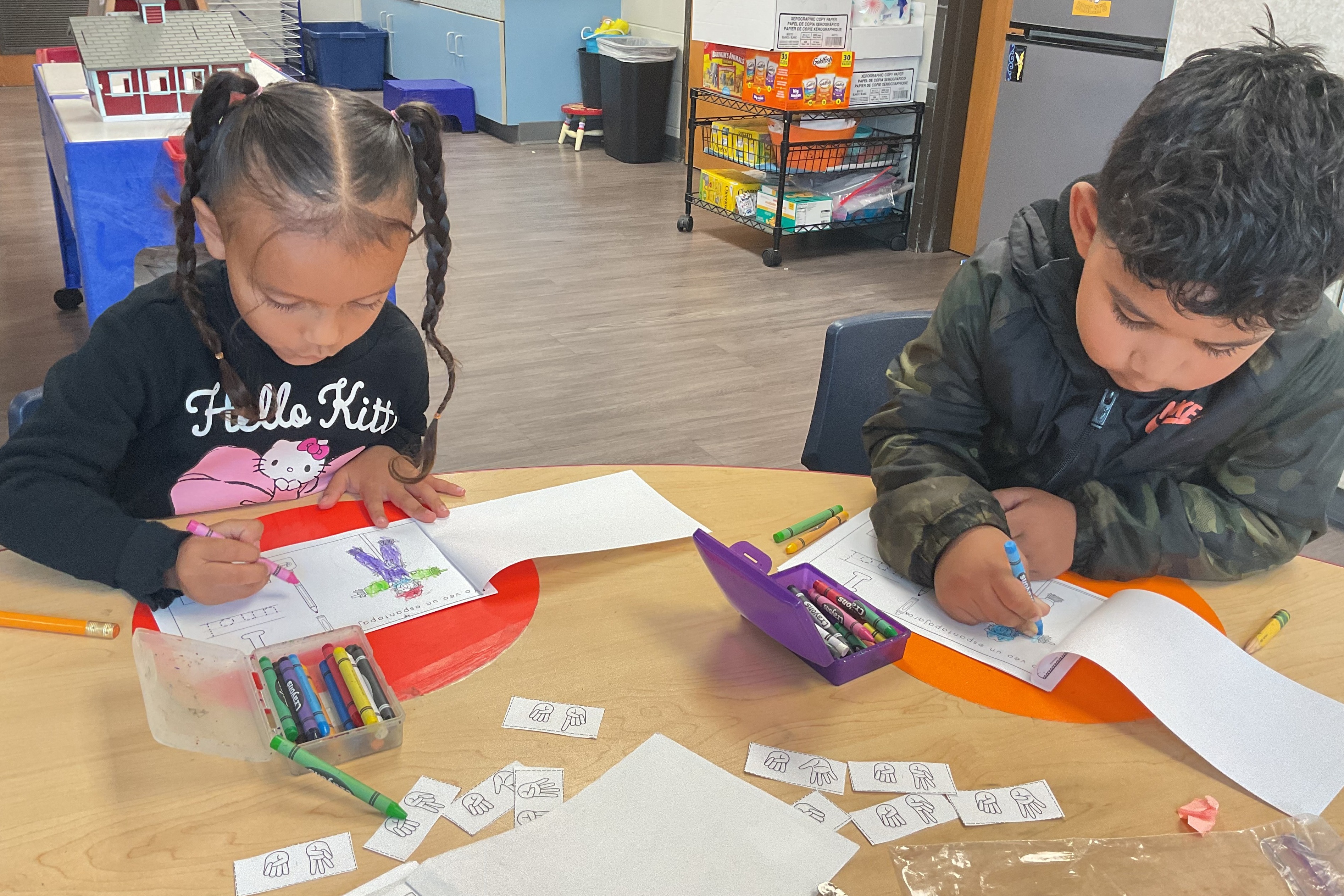 Two children work on paper and crayons at a table. One child wears a black shirt, the other a camouflage jacket.