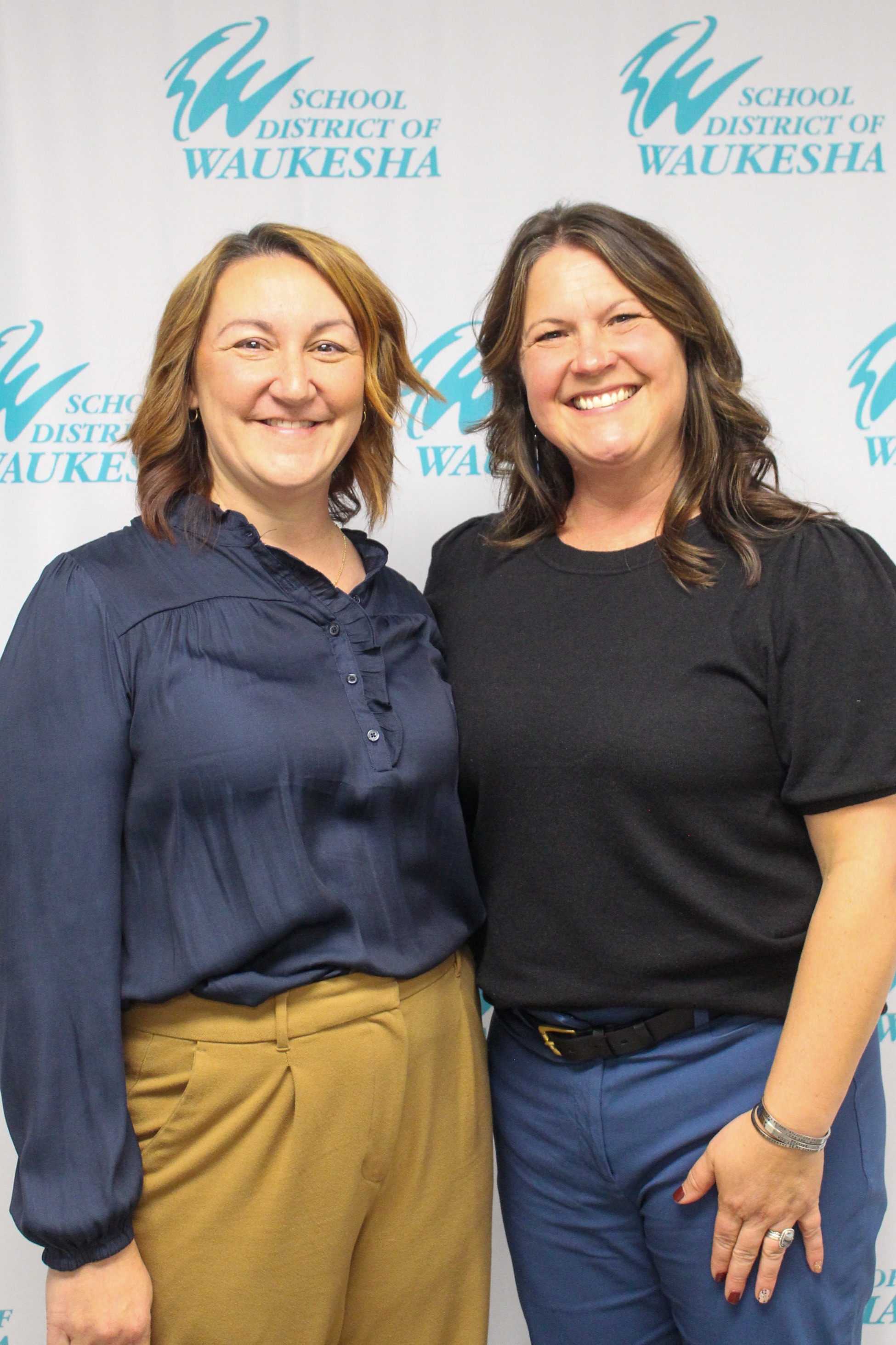 Two women smile, one in a blue blouse and khaki pants, the other in black with blue pants. Behind them, a white backdrop displays "School District of Waukesha" in blue.
