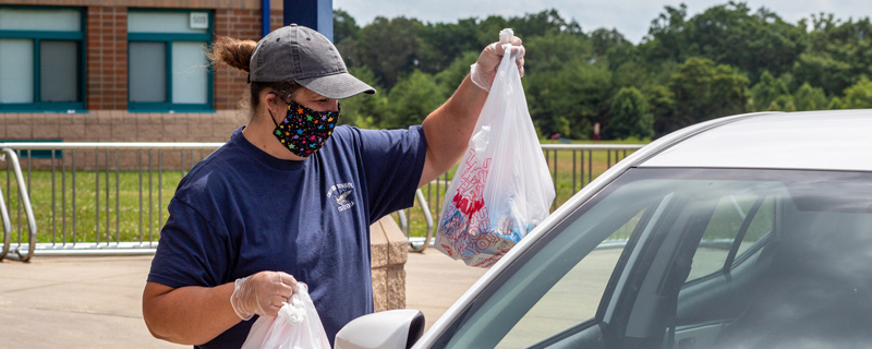 A masked staff member bringing a plastic bag with food to a car