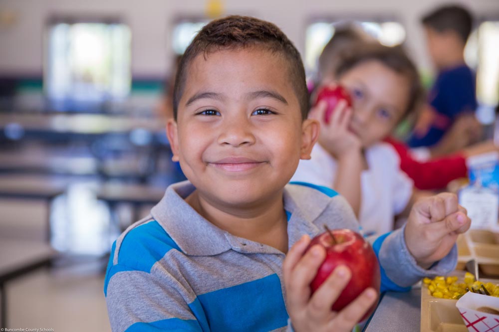 A child in the cafeteria holding an applet and smiling for the camera