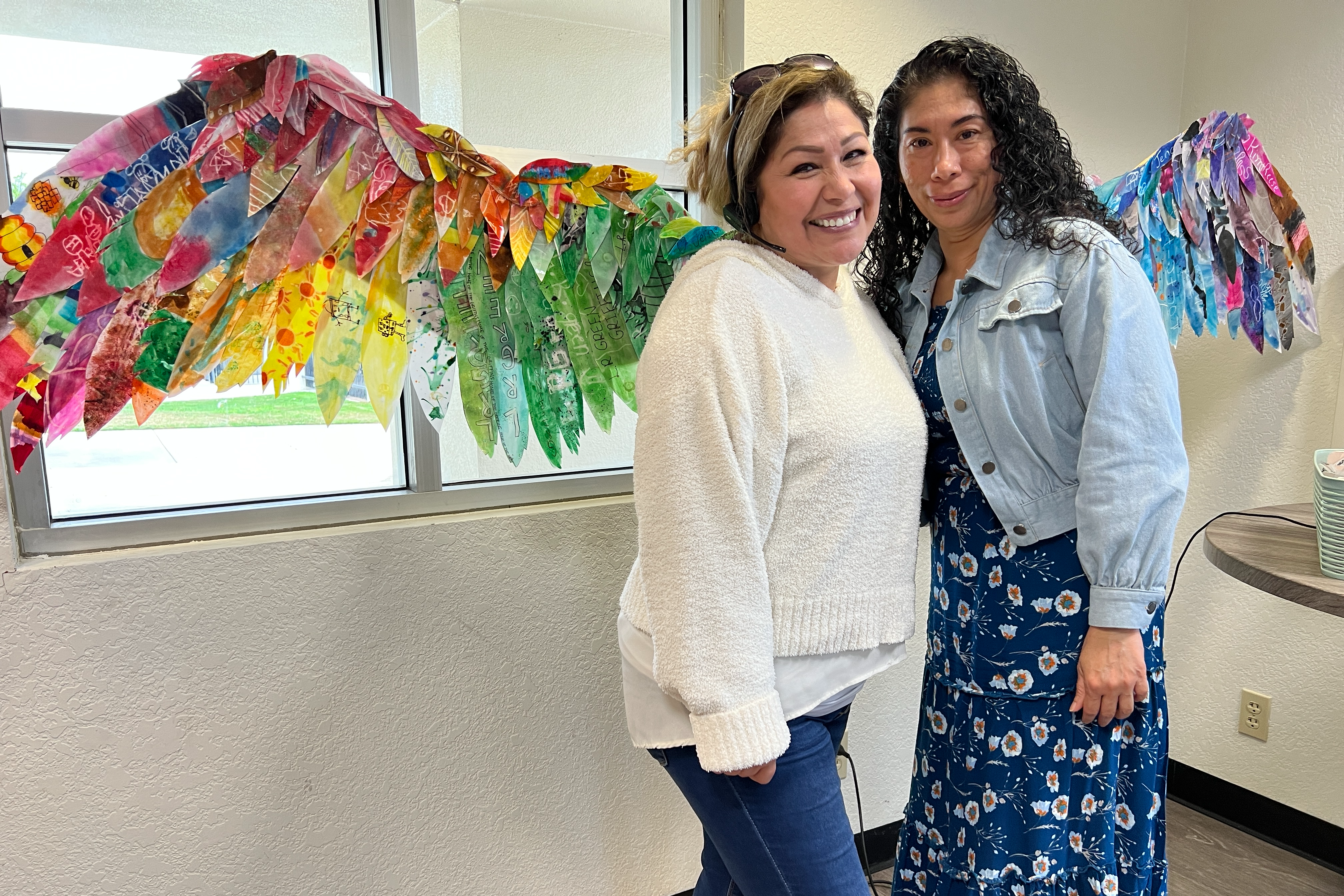 Two women smiling in front of colorful wings. 