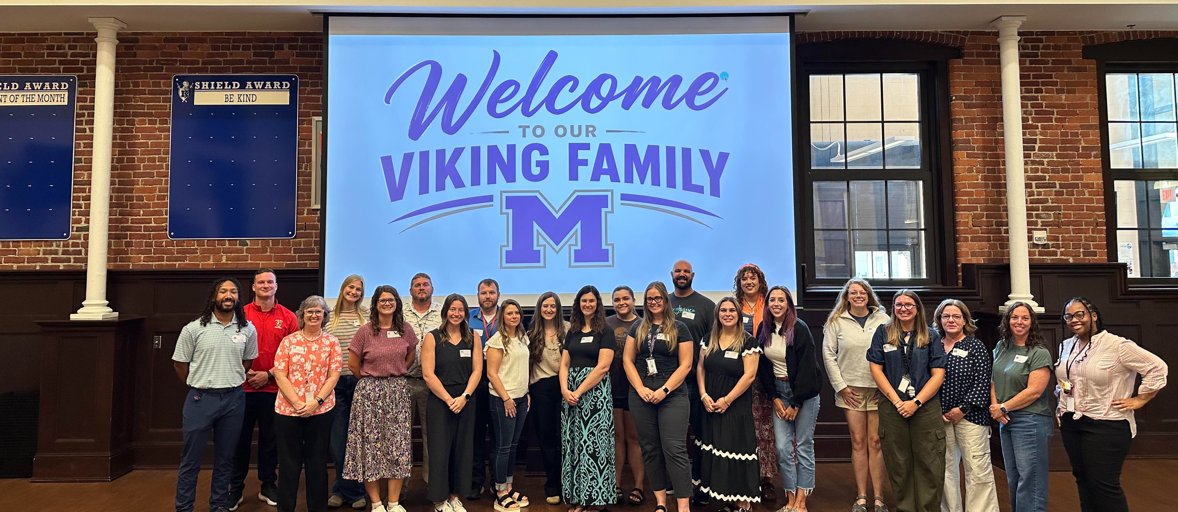 Group of people stand in front of a screen displaying "Welcome Viking Family" on a brick wall.