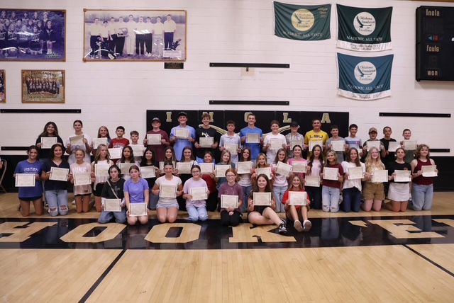 A group of students holding certificates, posing in a gymnasium with a wooden floor and banners.