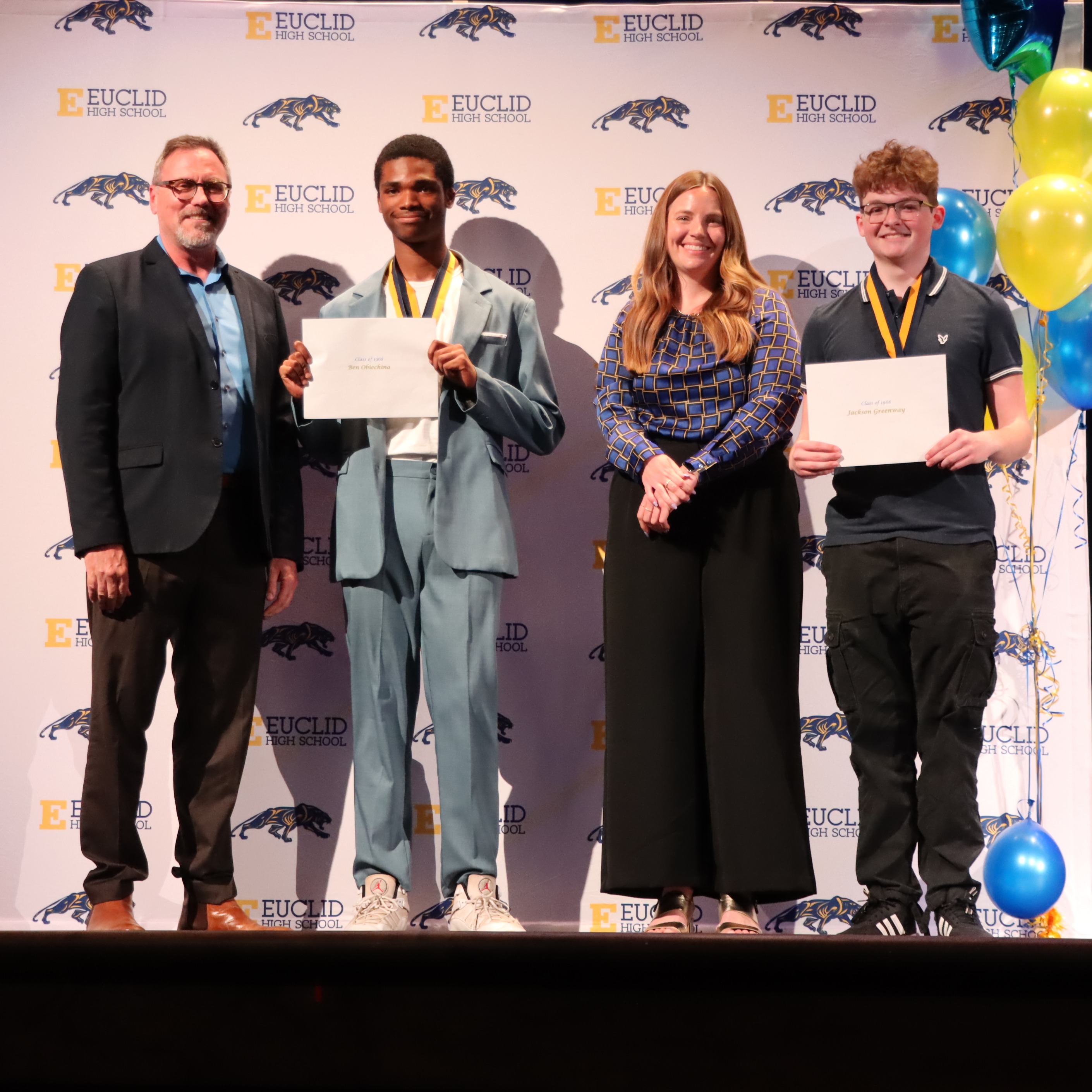 Two students stand in front of a Euclid Schools backdrop with scholarship envelopes while accompanied by Euclid Schools Foundation Board Members.