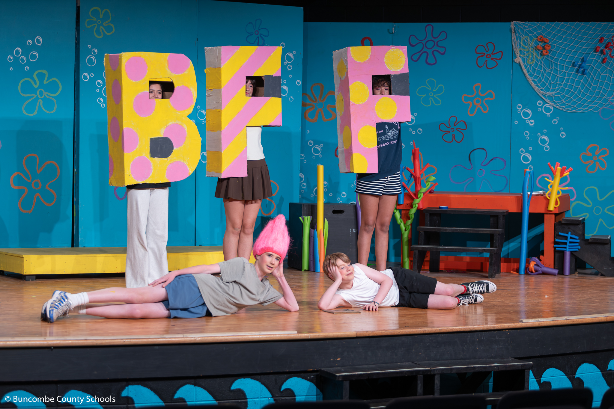 Student playing SpongeBob and Patrick Star lying on the floor of the stage. Behind them three students are wearing the letters B, F, and F. 