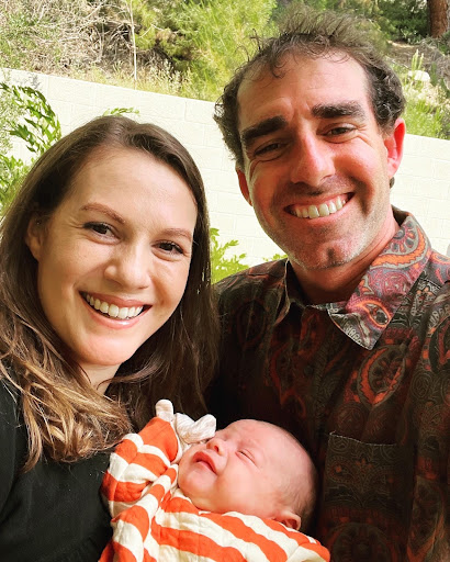 A couple cradles their newborn baby in their arms, with the man and woman both smiling down at the infant.