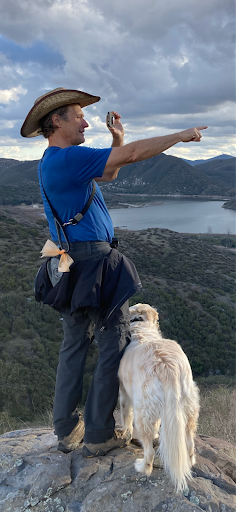 A male hiker stands triumphantly atop a rugged mountain peak, pointing up towards the sky while being accompanied by his dog.