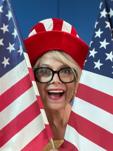 A woman wearing glasses and a top hat proudly holds two American flags.