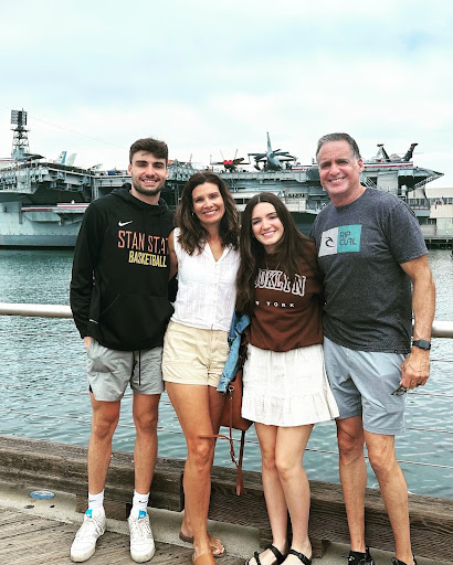 Two smiling men and two smiling women standing side by side together in front of an aircraft carrier docked at a pier.