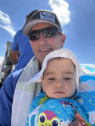 A man dressed in a blue t-shirt and sunglasses, displaying affection by holding an infant in his arms underneath a blue sky.