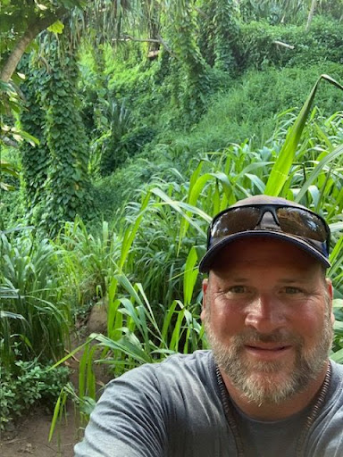 A man wearing a hat and sunglasses takes a selfie in front of a lush jungle.