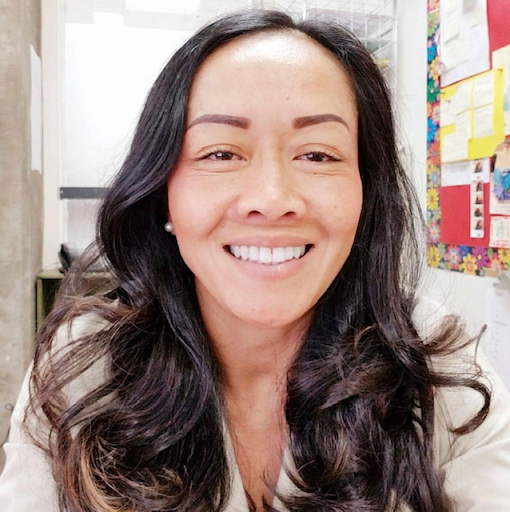 A smiling woman with long dark hair in a classroom environment looks directly at the camera.