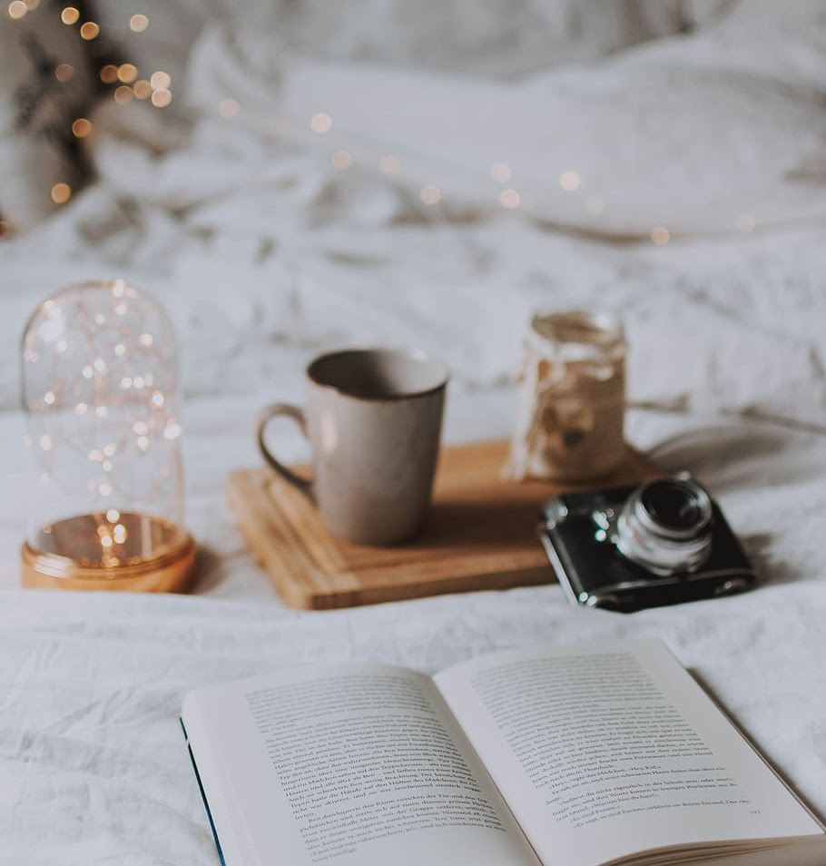 Cup, camera, christmas lights and open book on a white bed cover