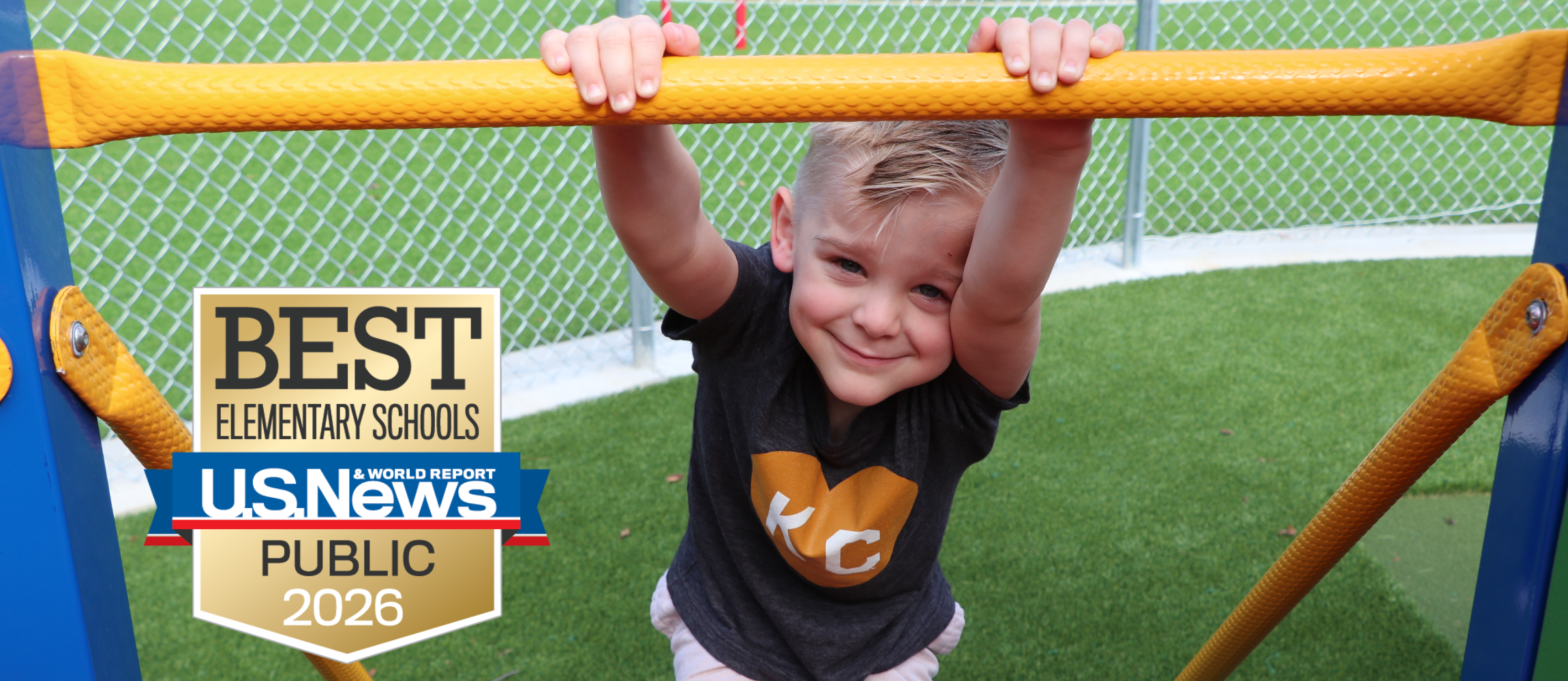 A preschool boy hangs on a bar of the playground equipment. There is a logo for the US News and World Report Best Elementary Schools logo in the bottom left.