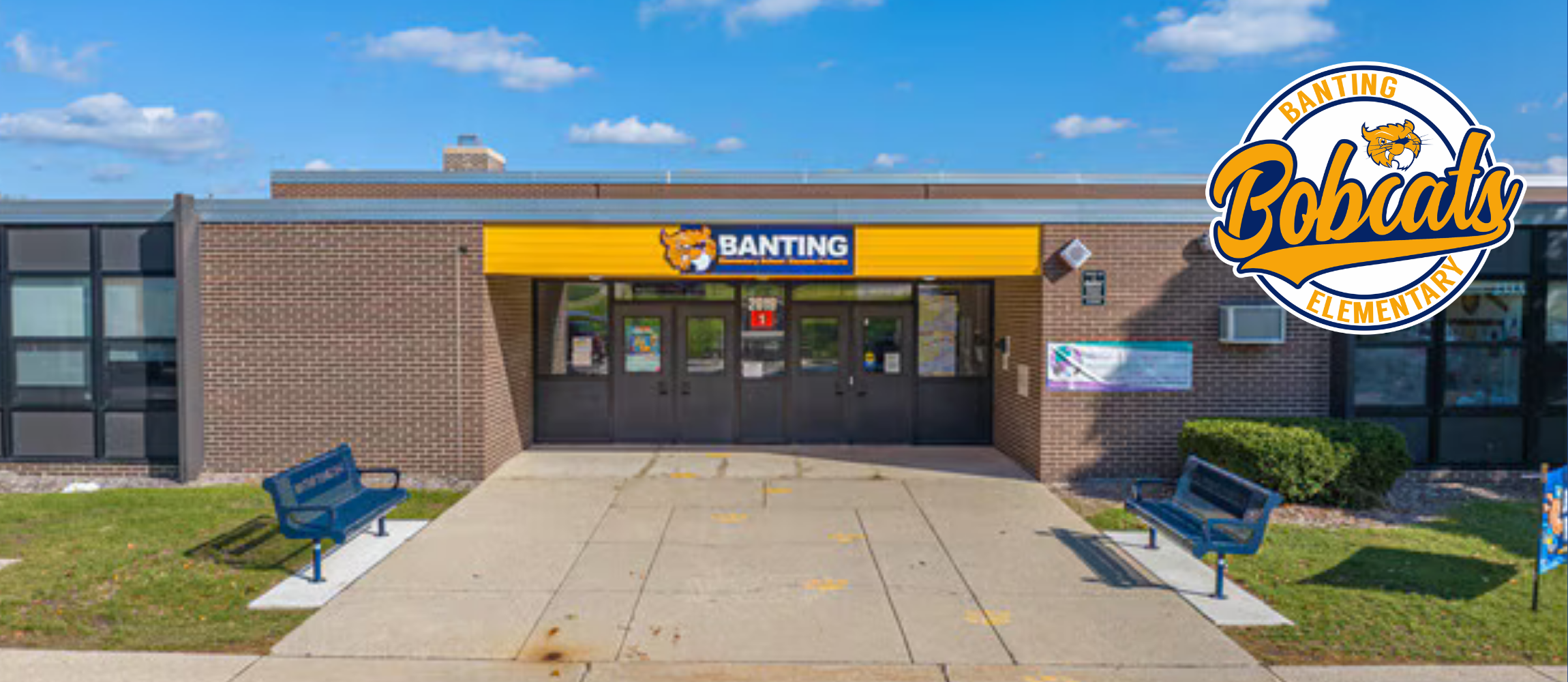 A building entrance with a yellow awning and logo. Two benches sit on either side of the entrance.