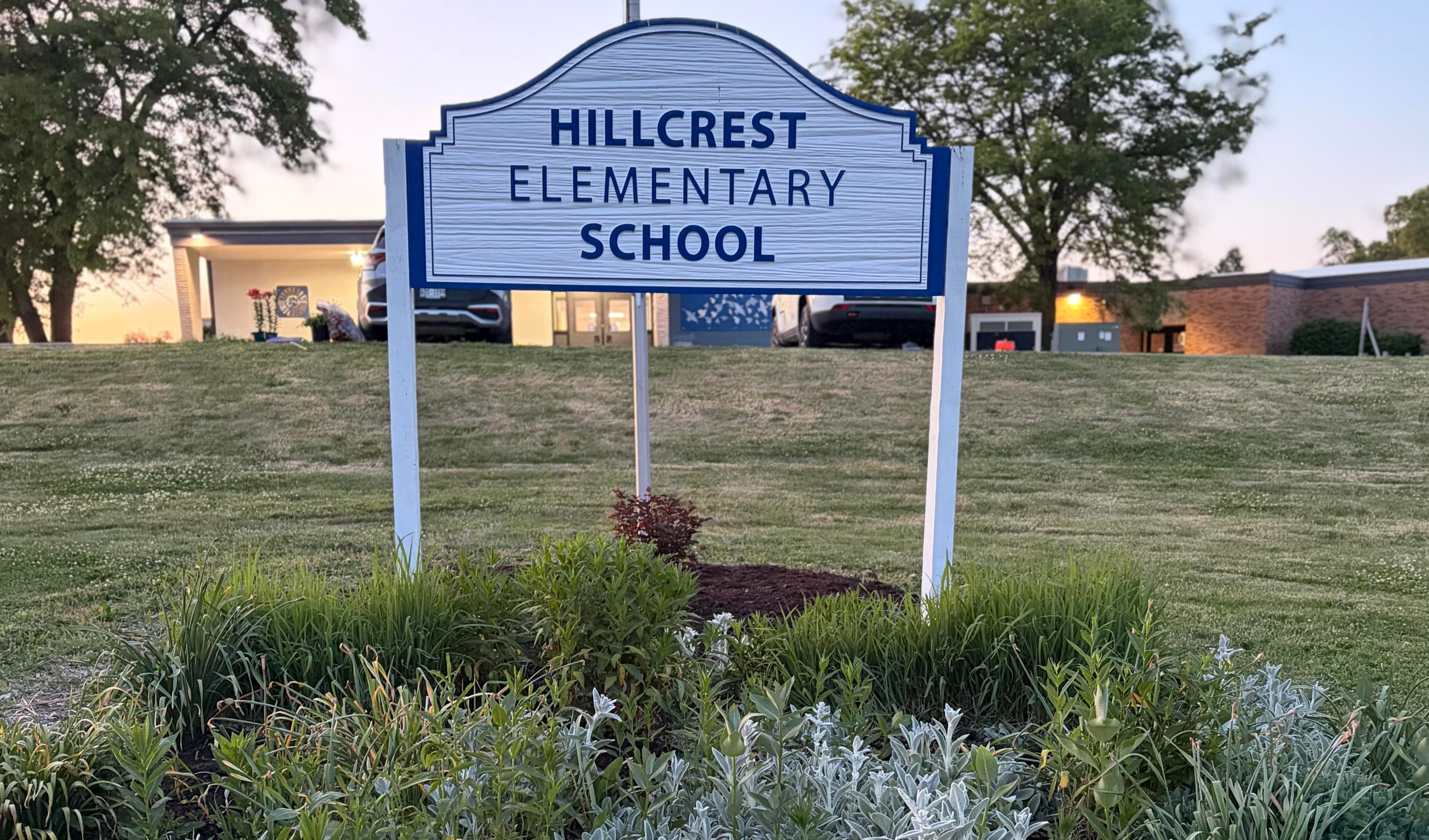 A sign for Hillcrest Elementary School stands before a garden. The sign has a white background with blue lettering.