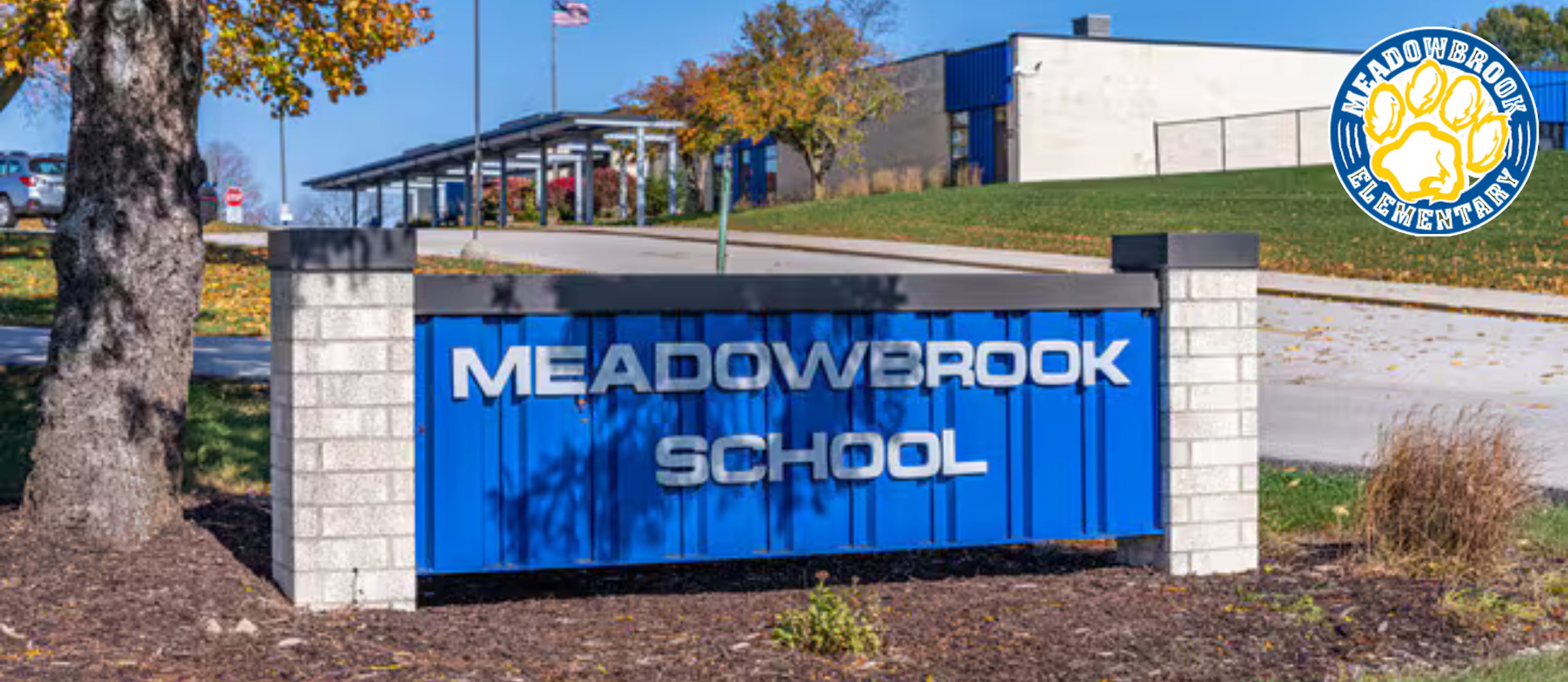 A blue Meadowbrook School sign stands before a grassy field with trees and a building.