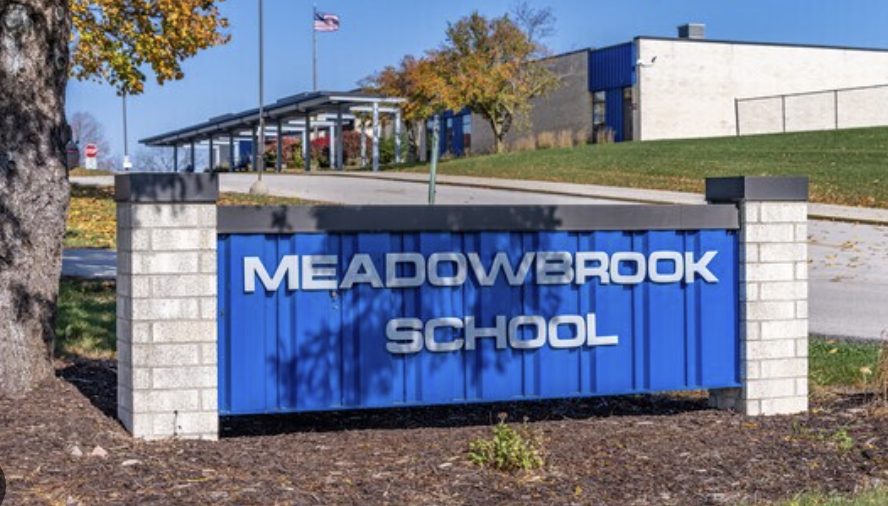 Blue sign with white text reads "MEADOWBROOK SCHOOL" against a grassy background with trees and a building.