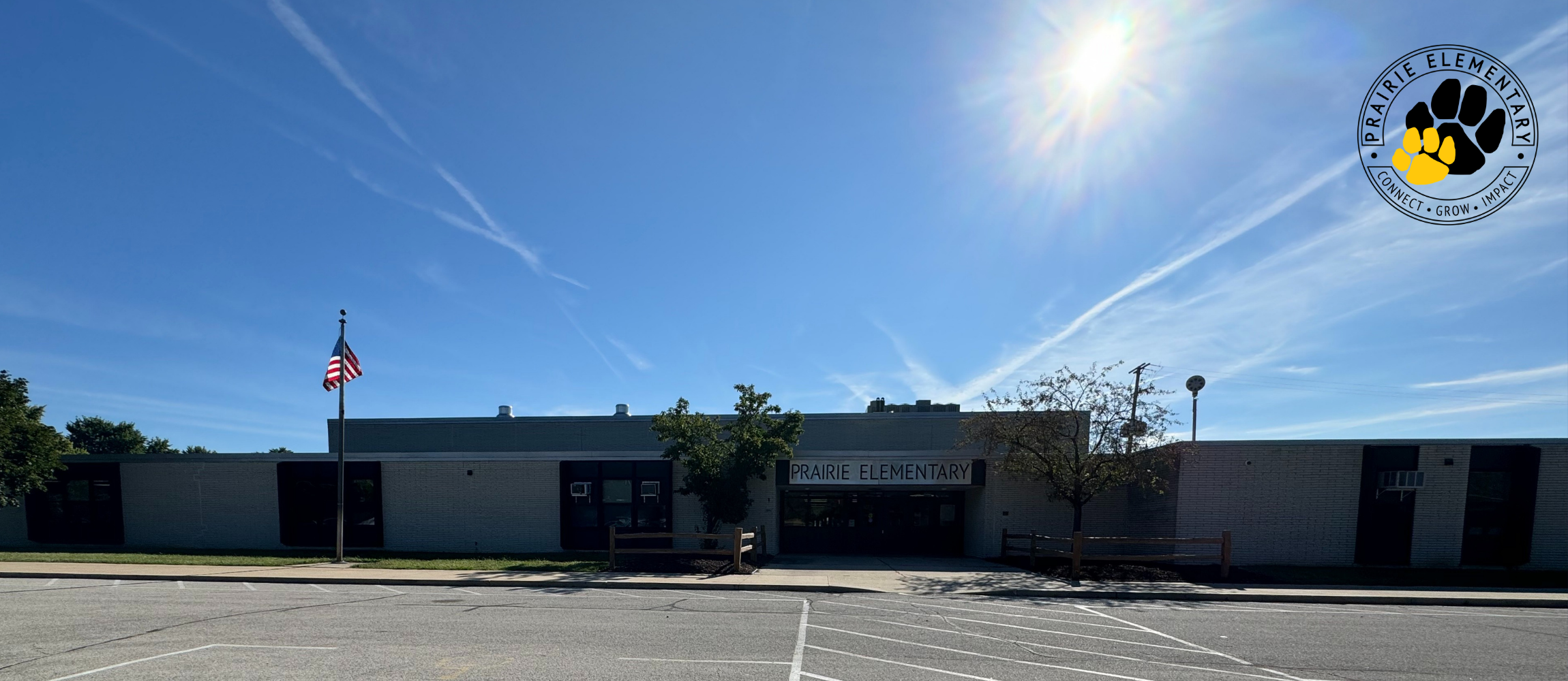 A school building with a flag pole, bright sunlight, and a clear blue sky. The road is empty.