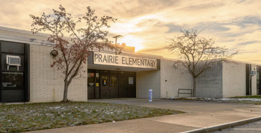 A school entrance with a sign reading "Prairie Elementary" on a clear, partly cloudy day. Trees and grass in front.