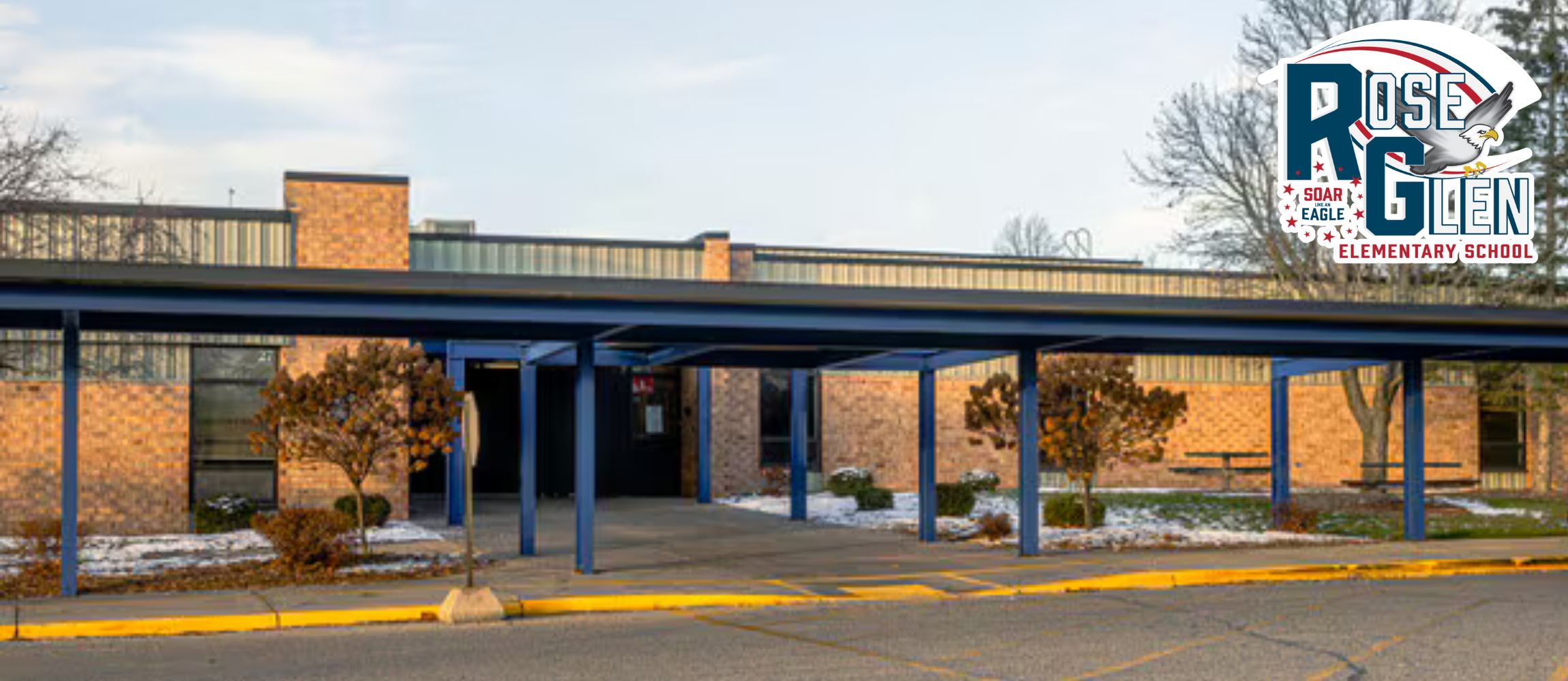 An elementary school building with a covered walkway and blue pillars, surrounded by snow and trees.