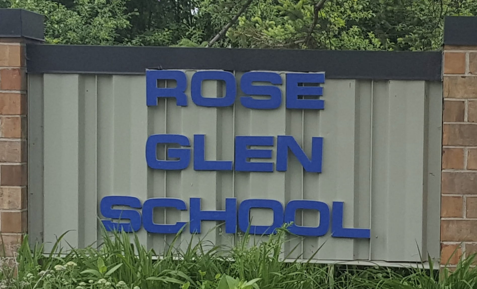 A gray sign with blue lettering reads "ROSE GLEN SCHOOL" against a background of brick walls and greenery.
