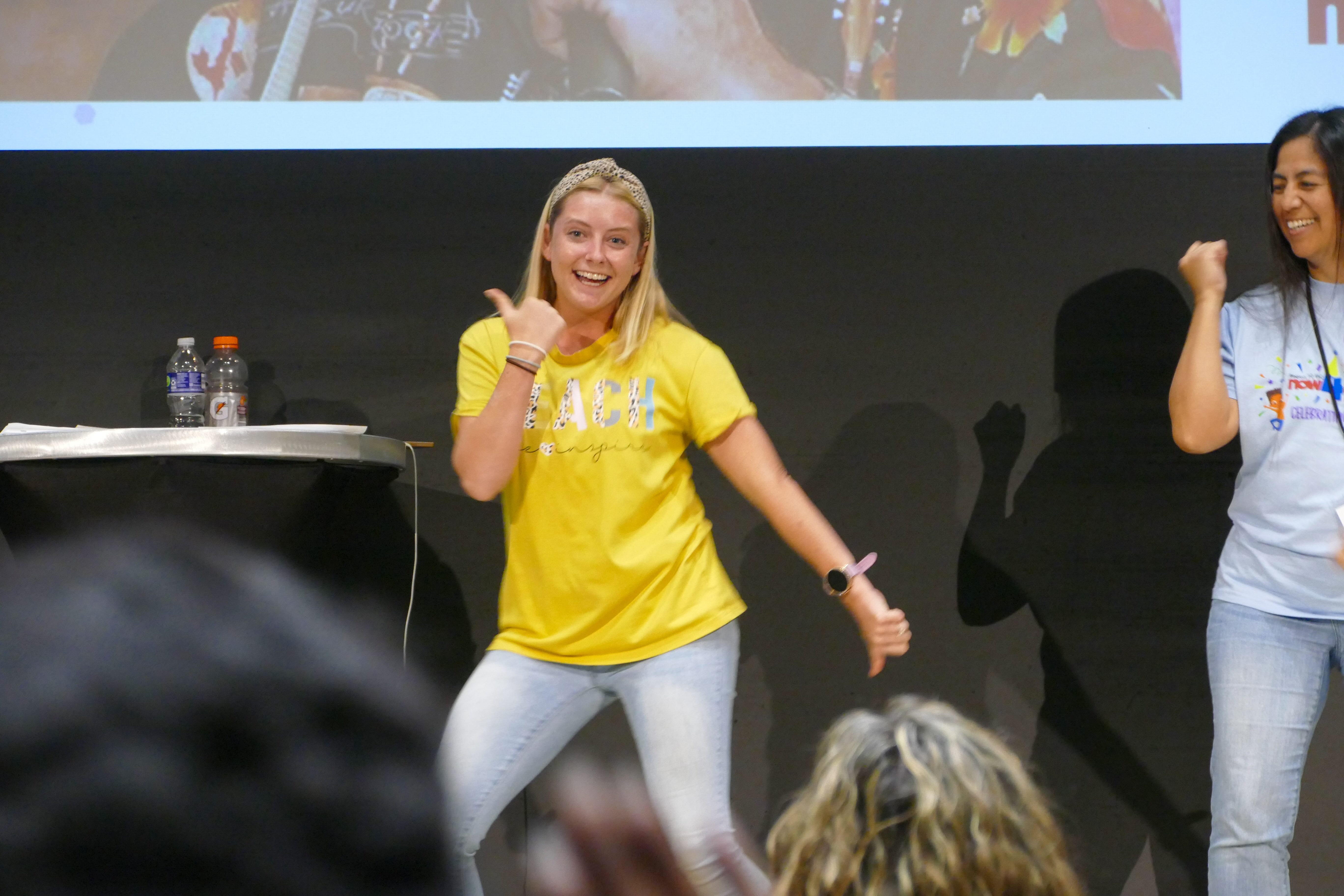 an educator wearing a yellow t-shirt and jeans smiles while swinging her arms for a dance. She is up on top of a stage.