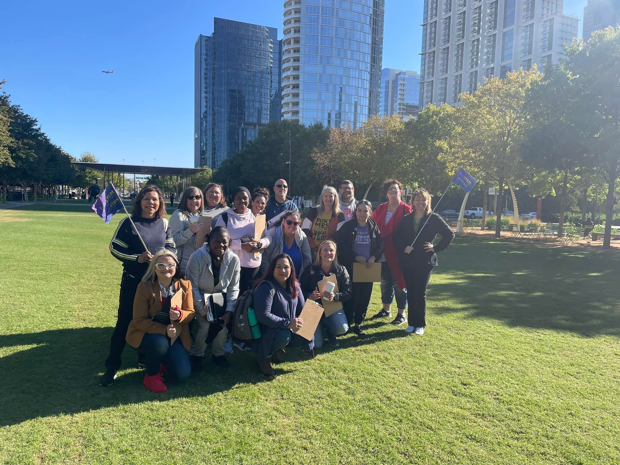 A group of people pose for a photo at on a green lawn in downtown Dallas. It is a sunny day, they are surrounded by skyscrapers.