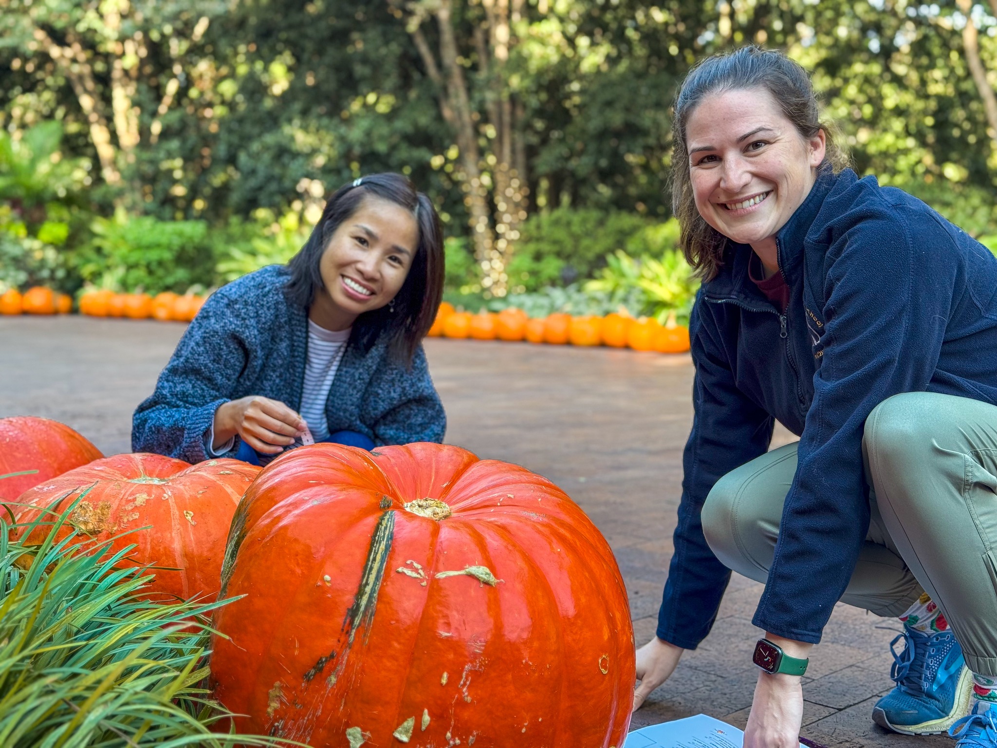 Two women pose for a photo. They are each next to a large orange pumpkin. They are outside in a beautiful garden with plenty of trees and greenery in the background.